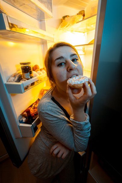Πορτραίτο γυναίκας δάγκωμα ντόνατ τη νύχτα κοντά σε ψυγείο - Φωτογραφία, εικόνα
