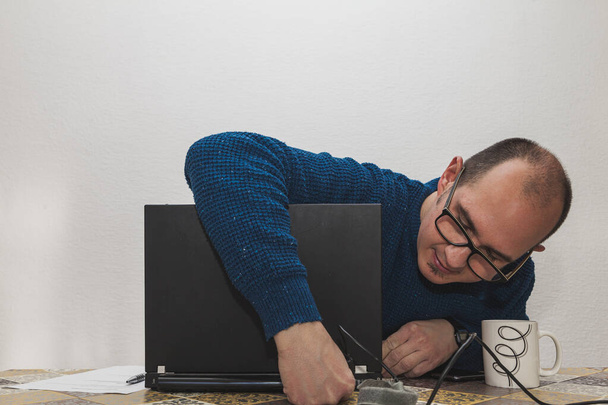 Un hombre con gafas y un suéter azul está conectando el cable de alimentación de un ordenador portátil. En la mesa hay una taza blanca, un smartphone, una hoja de papel y un bolígrafo. - Foto, imagen
