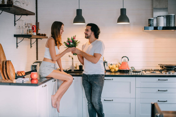 Día Internacional de la Mujer. Pareja joven por la mañana en la cocina en casa. Un hombre le da a una mujer un ramo de tulipanes el 8 de marzo en una hermosa cocina elegante. - Foto, imagen