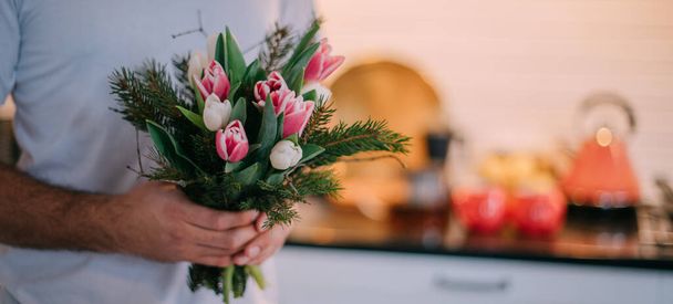 Męskie ręce z bukietem w kuchni. Międzynarodowy Dzień Kobiet. Młody facet trzyma w rękach bukiet tulipanów 8 marca. Romantyczny rodzinny wieczór w domu. - Zdjęcie, obraz