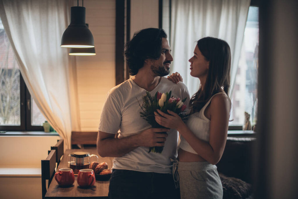 Casal amoroso à noite na sala de estar em casa. Dia Internacional da Mulher. Um homem dá a uma mulher um buquê de tulipas em 8 de março. Noite de família romântica em casa. - Foto, Imagem