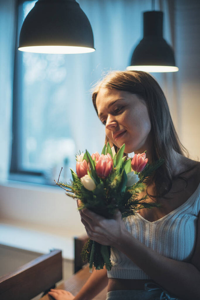 Portrait of a young woman with a bouquet of flowers. A beautiful girl sits on a table under the evening light of lamps with tulips in her hands. Sensual portrait in subdued light - Zdjęcie, obraz