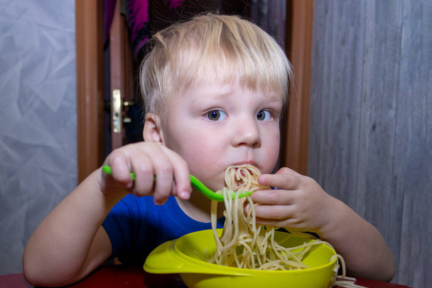 Ένα μικρό παιδί τρώει νουντλς στην κουζίνα, βοηθώντας τον εαυτό του με τα χέρια του. - Φωτογραφία, εικόνα