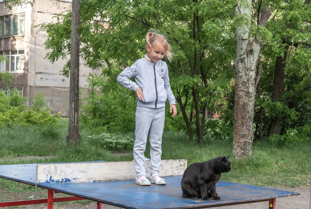 une petite fille, une européenne de cinq ans, se tient sur une table de tennis dans un parc, et un chat de rue noir s'assoit à côté d'elle - Photo, image
