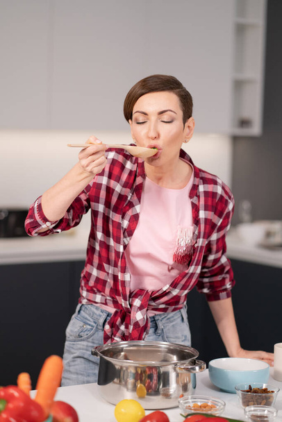 Pretty young housewife with a short hairs cooking a dinner for family trying it using a long wooden spoon standing in the modern kitchen. Healthy food at home. Close up - Foto, afbeelding