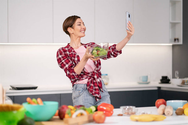 Pretty woman happy taking selfie using her smartphone while cooking fresh salad wearing a plaid shirt with a bob hair style. Healthy food leaving - vegan concept - Φωτογραφία, εικόνα