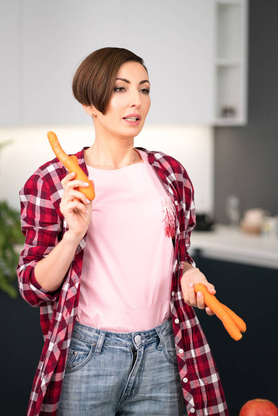 Charming short hair housewife with fresh carrot in the hands prepares carrot cake in the kitchen. Healthy food at home. Healthy food leaving concept - Фото, изображение