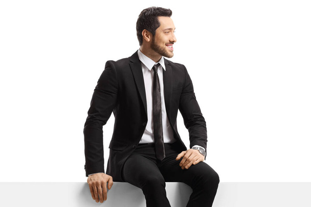 Young handsome man in a suit sitting on a panel isolated on white background - Φωτογραφία, εικόνα