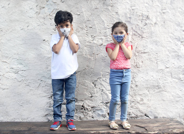 Bambini latini di 6 anni con maschere facciali covid-19, seduti con un muro bianco sullo sfondo, nuova normalità - Foto, immagini