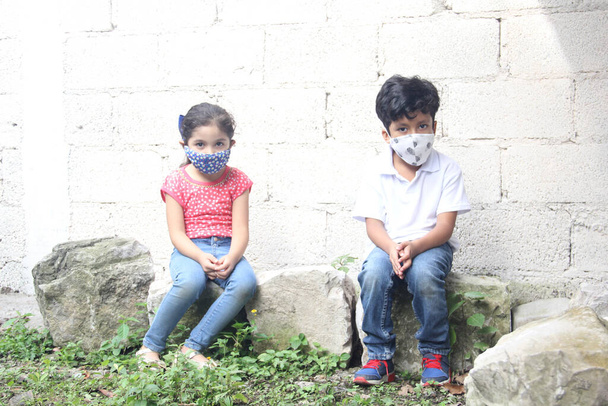 Niños latinos de 6 años con máscaras faciales covid-19, sentados con una pared blanca en el fondo, nueva normalidad - Foto, Imagen