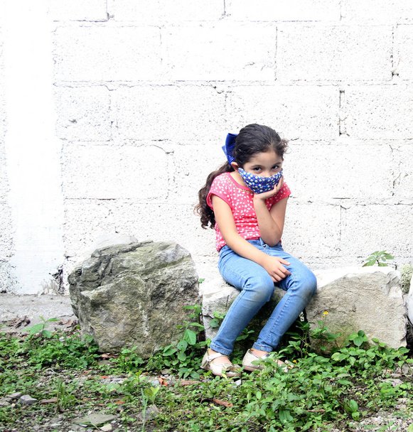 Fille latine de 6 ans avec masque de protection covid-19, assise sur des pierres avec un fond blanc, nouvelle normale dans les parcs - Photo, image