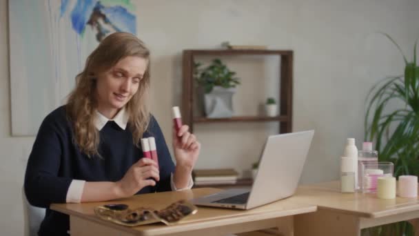 Keskikokoinen kuva nuoresta valkoihoisesta meikkitaiteilijasta istuu pöydän ääressä kannettavan tietokoneen edessä, jolla on videokeskustelu, jossa on kirkkaita huulipunia ja puhuu - Materiaali, video