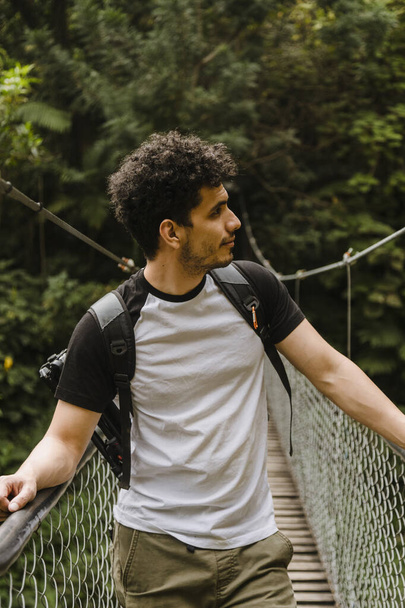 Portrait of young Hispanic man walking on a suspension bridge in the middle of the forest-Young traveler walking while he observes nature and enjoys the ride-Adventurous man - Photo, Image