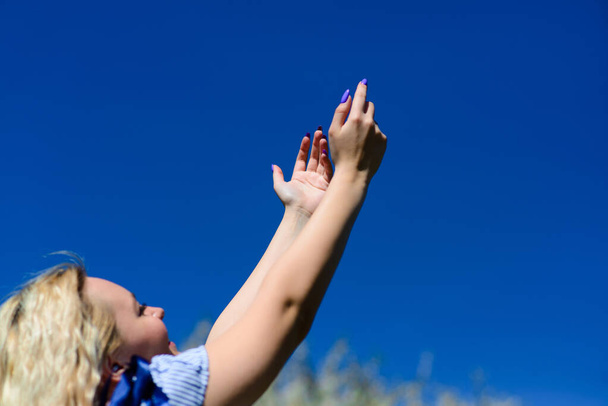 Μια νεαρή ξανθιά κοπέλα κοιτάζει τον όμορφο γαλάζιο ουρανό και φτάνει στον ουρανό με τα χέρια της. Όμορφη γυναίκα θαυμάζει τη φύση. - Φωτογραφία, εικόνα