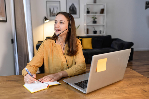 A young woman sits at the desk, using hands free headset and laptop for remote work, talking online and taking notes. Female call center operator works remotely - Φωτογραφία, εικόνα