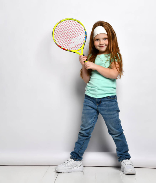 Beautiful smiling red-haired girl child big tennis player full length studio portrait. Little child in trendy outfit with racket posing for camera. Kids sport motivation, enjoyment active lifestyle - 写真・画像
