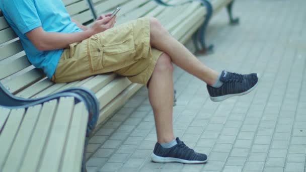 Hombre con teléfono móvil se sienta con las piernas cruzadas en el banco de la ciudad, seguimiento de la cámara - Metraje, vídeo
