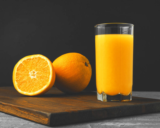 Glass of fresh orange juice, whole and sliced fruits on wooden board on dark background with copy-space. - Foto, Imagem