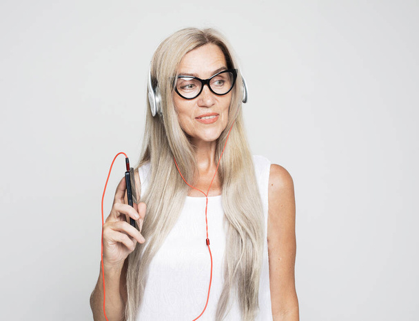 Old people, emotion and technology concept: portrait of happy senior woman with long white hair wearing eyeglasses listening to music with headphones over grey background - Photo, Image