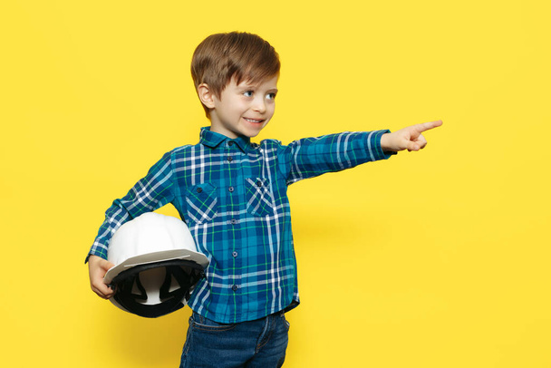 Happy little kid boy engineer or architect in a protective helmet pointing finger away over yellow background. - Fotoğraf, Görsel