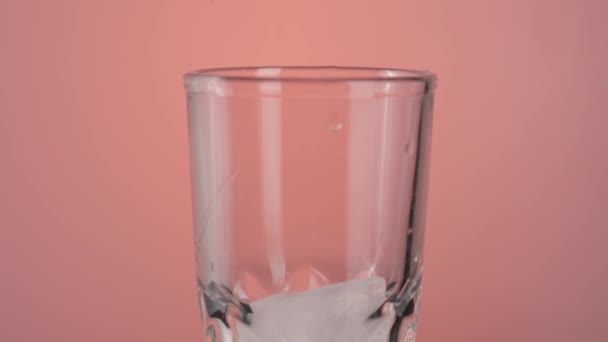 Primo piano di cubetti di ghiaccio che cadono in un bicchiere vuoto cocktail isolato su sfondo rosa. Azione. Preparare un cocktail con ghiaccio. - Filmati, video