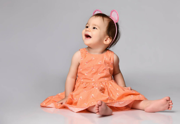 Portrait of a sweet baby girl dressed in a dress and a hoop with ears sitting on gray in the studio. - Foto, afbeelding