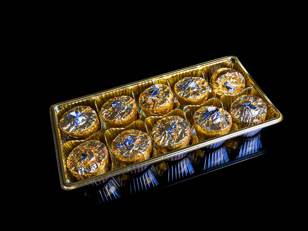 Een doos met ronde snoepjes in gouden folie op een zwarte achtergrond. Verpakte bonbons. Ronde taart. Lekker eten. Een mooi cadeau. Gouden kleur. Voedselfoto. Product. Delicatie. Achtergrondbeeld. - Foto, afbeelding