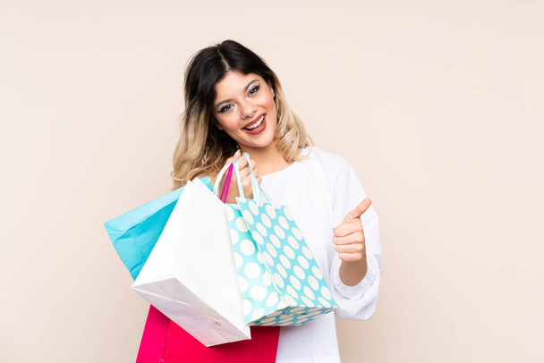 Teenager girl isolated on beige background holding shopping bags and with thumb up - Fotografie, Obrázek