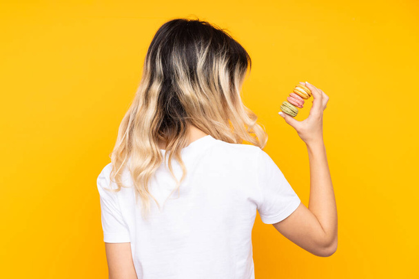 Menina adolescente isolada no fundo amarelo segurando macarons franceses coloridos na posição traseira - Foto, Imagem