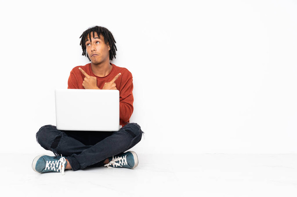 Mladý africký Američan sedí na podlaze a pracuje s laptopem ukazujícím na laterály, kteří pochybují - Fotografie, Obrázek