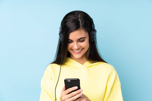 Jonge vrouw over geïsoleerde blauwe achtergrond luisteren muziek en op zoek naar mobiele - Foto, afbeelding