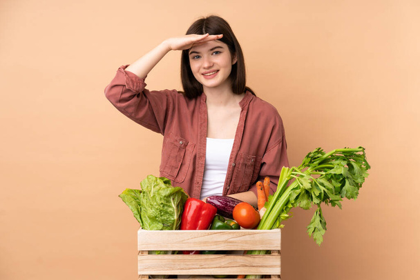 Jonge boer meisje met vers geplukte groenten in een doos kijken ver weg met de hand om iets te kijken - Foto, afbeelding