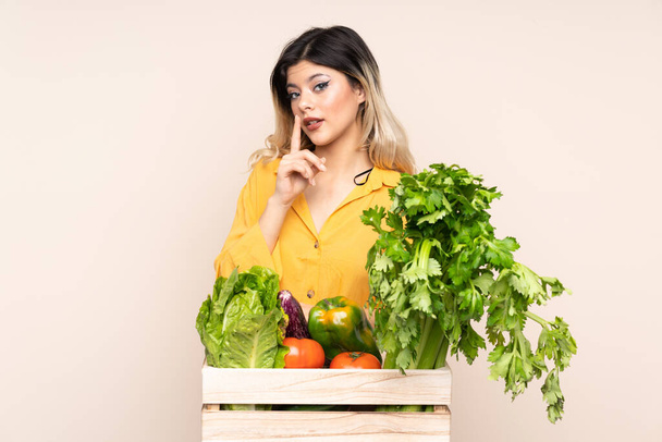 Ragazza contadina adolescente con verdure appena raccolte in una scatola isolata su sfondo beige facendo gesto di silenzio - Foto, immagini