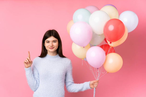 Молода українська дівчина-підліток, що тримає багато повітряних кульок над ізольованим рожевим фоном, показує з вказівним пальцем чудову ідею. - Фото, зображення