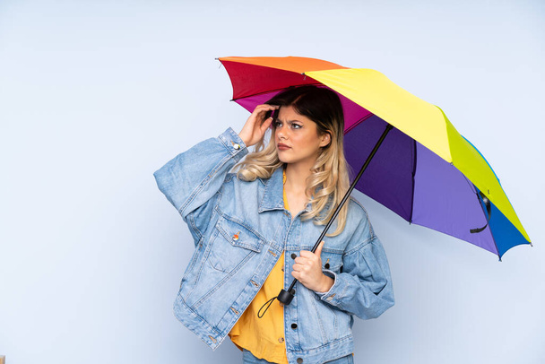 Teenager rusky dívka drží deštník izolovaný na modrém pozadí s pochybnostmi a s zmateným výrazem - Fotografie, Obrázek