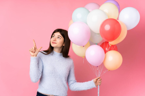 jong Oekraïense tiener meisje houden veel ballonnen over geïsoleerde roze achtergrond wijzend met de wijsvinger een geweldig idee - Foto, afbeelding