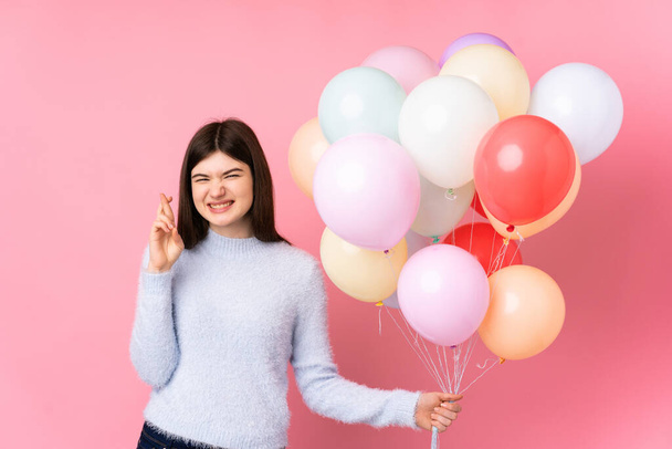 Young Ukrainian teenager girl holding lots of balloons over isolated pink background with fingers crossing - Fotografie, Obrázek
