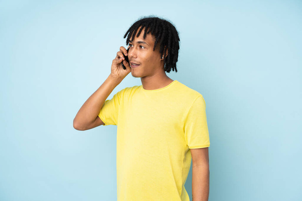 Młody Afroamerykanin odizolowany na niebieskim tle prowadzący rozmowę z telefonem komórkowym - Zdjęcie, obraz