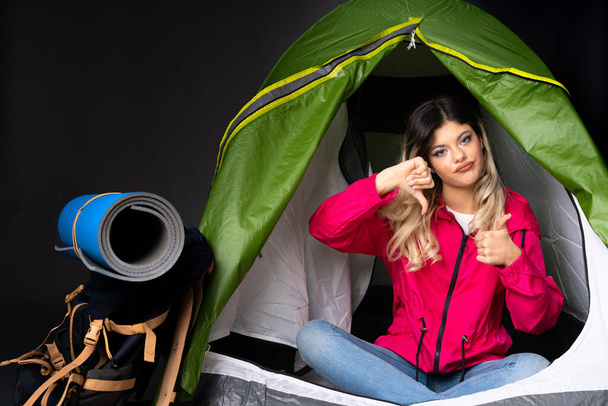Menina adolescente dentro de uma barraca verde acampamento isolado no fundo preto fazendo bom-mau sinal. Indeciso entre sim ou não - Foto, Imagem