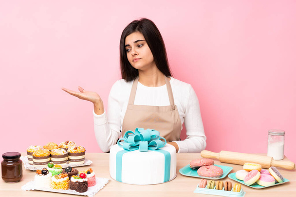 Pastry chef with a big cake in a table over isolated pink background holding copyspace with doubts - 写真・画像