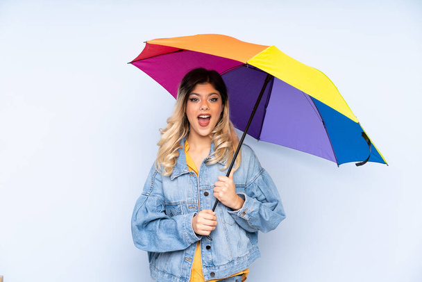 Teenager russian girl holding an umbrella isolated on blue background with surprise and shocked facial expression - Foto, Bild