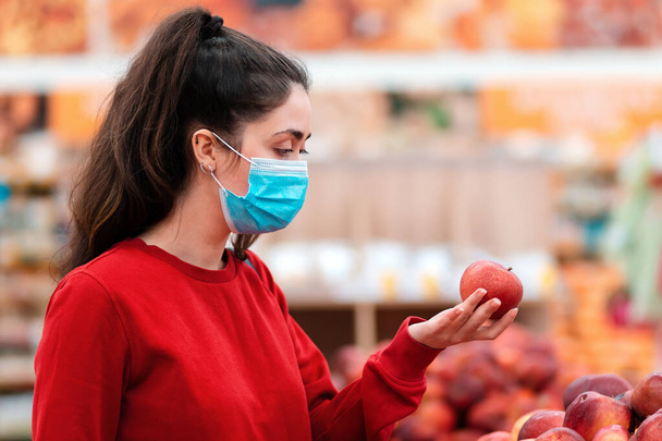 Einkaufen. Porträt einer jungen Frau mit medizinischer Maske auf dem Gesicht, die in einem Supermarkt Äpfel auswählt. Das Konzept des Konsumismus und die neue Realität. - Foto, Bild