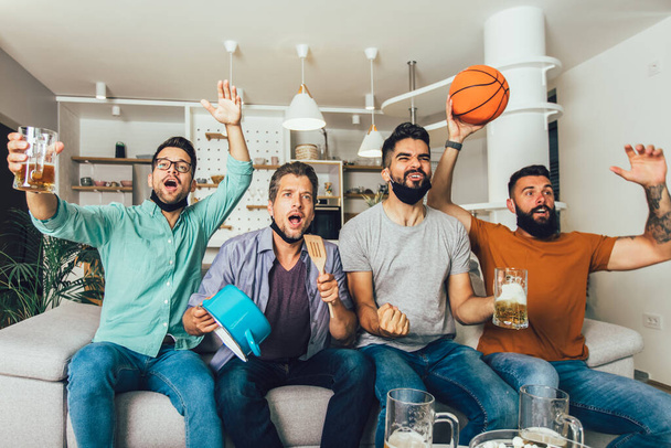 feliz masculino amigos vestindo preto rosto máscara protetora assistindo basquete em casa - Foto, Imagem