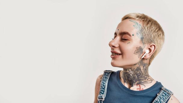 Listening to music. A close up of a white girl tattooed with piercing wearing denim overall standing and listening to music with her wireless earphones eyes closed and smiling - Zdjęcie, obraz