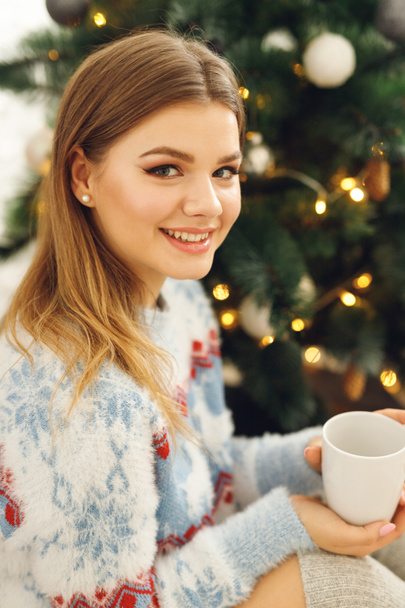 Close up of girl wears festive clothes take cup of coffee at home near Christmas Tree. Smiling woman sit on mat in blue sweater with deer and gray stockings - Foto, immagini
