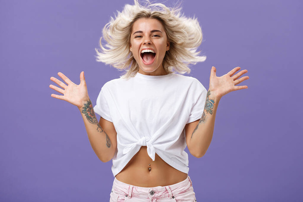 Studio shot of happy carefree and pleased joyful woman with blonde hair spreading palms aside in delighted pose smiling broadly and gazing entertained at camera jumping while having fun - Photo, Image