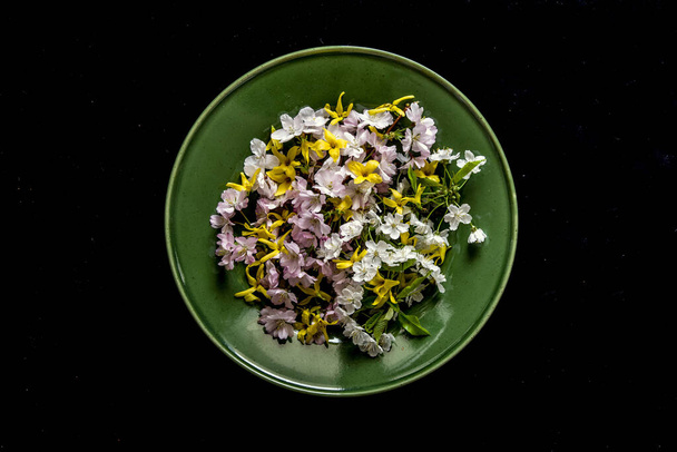 An overhead shot of small picked flowers on  green plate on a black surface - Φωτογραφία, εικόνα