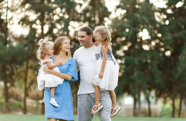 Rodiče drží dcery v náručí na venkově a užívají si společně. Šťastný rodinný koncept. Rozmazané stromy na pozadí. Perfektní rodina - Fotografie, Obrázek
