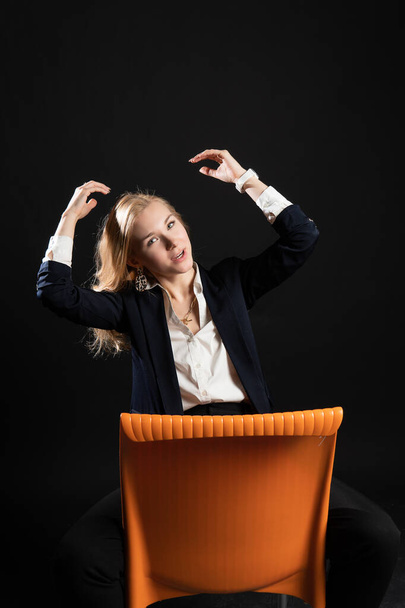 girl sitting on an orange chair. Cheerful looks at the camera, enjoy the concept, on a black background in the studio, in blazer business woman, blonde - Φωτογραφία, εικόνα