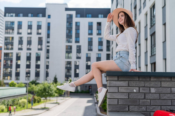 Beautiful young woman in shorts and a hat posing while sitting on a stone bench. A full-length portrait of a Caucasian girl on a hot summer day. - Fotografie, Obrázek